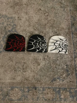 Mössa med spindelnätsmönster - Tre mössor i olika färger med coolt spindelnätsmönster. En svart med rött nät, en svart med vitt nät och en vit med svart nät. Perfekt accessoar för dig som gillar streetwear och vill sticka ut. Materialet ser ut att vara mjukt och stretchigt, troligen bomull eller akryl.