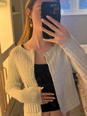 Ljusgrå kabelstickad kofta - Säljer en ljusgrå kabelstickad kofta med rund hals och knappar framtill. Koftan har långa ärmar och ribbade muddar, perfekt för lager på lager. Klassisk och enkel design som passar till det mesta.