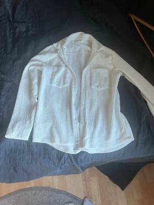 Vit ribbad skjorta med fickor - Snygg vit skjorta med ribbad struktur och två bröstfickor. Skjortan har knappar framtill, klassisk krage och lång ärm. Perfekt för en avslappnad men ändå stilren look. Även perfekt till sena sommarkvällar💓💓 använd noll gånger💓vet ej varumärket 
