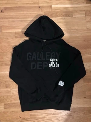 Svart Gallery Dept Hoodie - Svart hoodie från Gallery Dept med stor huva och dragsko. Framsidan har trycket 'GALLERY DEPT.' och 'DÉPT. de la GALERIE' i vitt och svart. Baksidan har ett stort vitt emblem. Tillverkad i mjuk bomull, perfekt för en avslappnad streetwear-look.