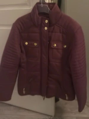Vinröd pufferjacka med gulddetaljer - Säljer en vinröd pufferjacka med hög krage och guldfärgade knappar och dragkedja. Jackan har två bröstfickor med lock och dekorativa knappar samt två sidofickor med dragkedja. Perfekt för kalla vinterdagar och riktigt snygg design.