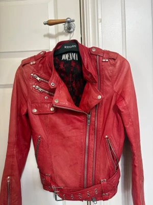 Röd skinnjacka från Jofama - Snygg röd skinnjacka från Jofama med coola detaljer som asymmetrisk dragkedja, flera fickor med dragkedjor, axelklaffar och bälte i midjan. Jackan har en bikerstil och silverfärgade metalldetaljer som ger en edgy look.