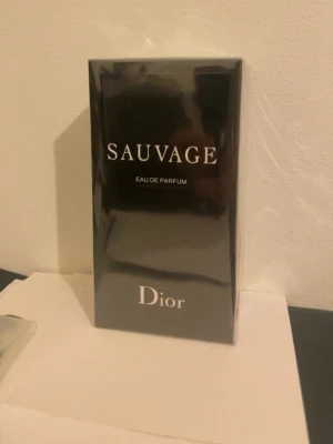 Dior Sauvage Eau de Parfum - Dior Sauvage Eau de Parfum i elegant svart förpackning med vit text. Kartongen har en stilren och lyxig design med märkets logotyp på toppen. Perfekt för dig som gillar exklusiva dofter och vill ha något klassiskt på parfymhyllan.