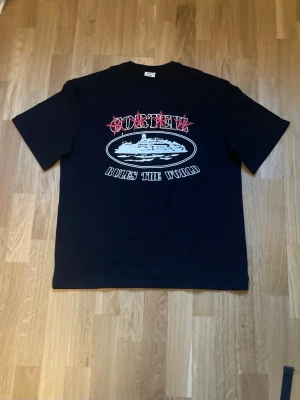 Svart t-shirt corteiz Rules The World - Svart t-shirt från corteiz med tryck framtill som visar ett fartyg och texten 'Rules The World'. T-shirten har klassisk passform och rund hals. Perfekt för dig som gillar streetwear och grafiska prints.