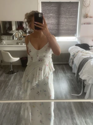 Vit blommig långklänning med volang - Jätte fin vit långklänning, tunna axelband, blommigt mönster i pastellfärger och volang detaljer. Perfekt för varma dagar och speciella tillfällen som tillexempel midsommar. Använd 1 gång. Nypris 600.