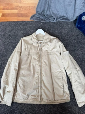 Beige vindjacka från Stone Island - Snygg beige vindjacka från Stone Island med klassisk patch på ärmen. Jackan har dragkedja framtill, raka ärmar och enkel krage. Tillverkad i lätt och vindtätt material som passar perfekt för blåsiga dagar. Minimalistisk design och diskreta detaljer.