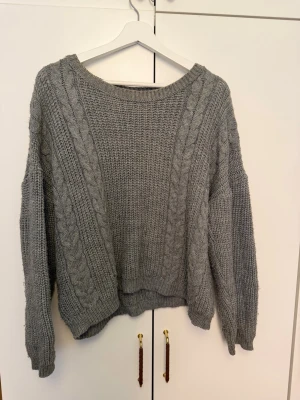 Grå stickad tröja från First Class - Mysig grå stickad tröja från First Class i storlek S (36-38). Tröjan har ett klassiskt flätmönster framtill, rund halsringning och långa ärmar. Perfekt för kyliga dagar och enkel att matcha med jeans eller kjol.