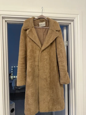 Beige teddyjacka från Stand Studio - Säljer en beige teddyjacka från Stand Studio med klassisk krage och mjukt, fluffigt material. Jackan är lång och har en rak passform som ger en snygg siluett. Perfekt för dig som vill ha en trendig och mysig jacka till hösten och vintern. Ena fickan har ett trasigt foder, det står st 50 men som en L