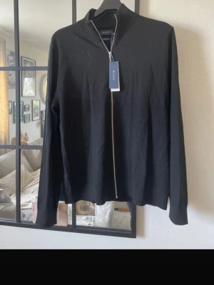 Svart merinoullströja från Riley - Stilren svart tröja från Riley i mjuk merinoull. Tröjan har hel dragkedja framtill, hög krage och långa ärmar. Perfekt för dig som gillar en clean och klassisk look med premiumkänsla. Helt ny