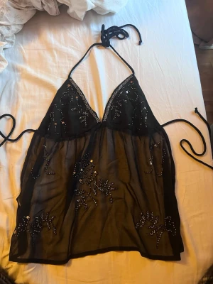 Svart transparent halterneck topp - Snygg svart transparent halterneck topp med knyt i nacken och ryggen. Toppen har v-ringning och är dekorerad med svarta paljetter i ett mönster framtill och baktill. Perfekt för dig som vill sticka ut lite extra.
