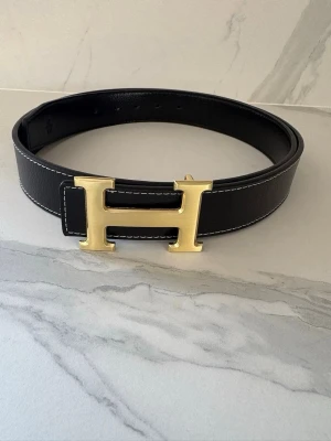 Svart H-bälte med gulddetalj - Snyggt svart skärp med en stor, ikonisk H-spänne i guld. Bältet är gjort i äkta skinn och har vita kontrastsömmar längs kanterna. Perfekt accessoar för att lyfta vilken outfit som helst.