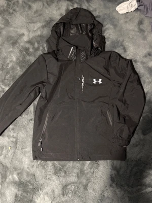 Svart vindjacka från Under Armour - Snygg svart vindjacka från Under Armour med huva och dragkedja framtill. Jackan har två sidofickor med dragkedja, justerbara muddar och logga på bröstet. Materialet är lätt och vattenavvisande, perfekt för blåsiga dagar.