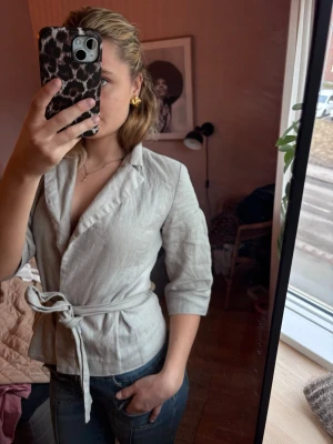 Ljusbeige linnekavaj med knytskärp - Snygg ljusbeige kavaj i linne med trekvartsärmar och knytskärp i midjan. Klassisk krage och figurnära passform som ger en stilren look. Perfekt att styla med jeans eller kjol för en trendig outfit.