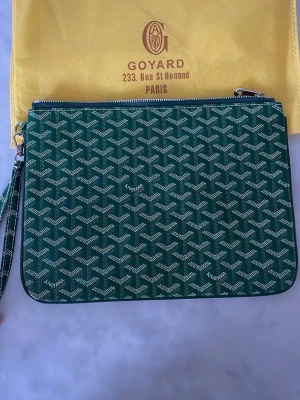 Plånbok och Ipadfodral Lindex - Snygg grön kuvertväska med vita mönstret och silverfärgad dragkedja. Väskan har en avtagbar handledsrem och insidan är fodrad i gult tyg med en praktisk innerficka. Perfekt för att bära det viktigaste med stil. Samt fet plånbok