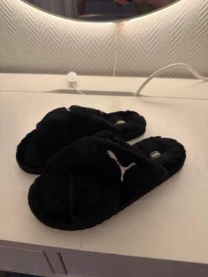 Svarta fluffiga tofflor från Puma - Mysiga svarta tofflor från Puma med korslagda band och broderad logga på ovansidan. Tillverkade i mjukt, fluffigt material som känns skönt mot fötterna. Perfekta för att chilla hemma och hålla värmen under kalla dagar.