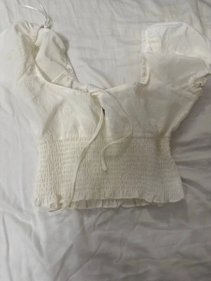 Vit blus med puffärm och knyt - Söt vit blus med puffärmar och knyt framtill. Toppen har smockad midja och volangkant nedtill för en romantisk look. Perfekt för dig som gillar feminina detaljer och en luftig känsla.