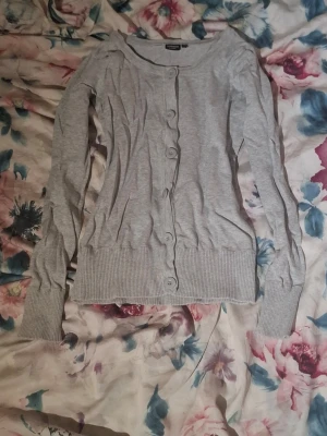 Ljusgrå kofta från kappAhl - Ljusgrå kofta från WeTweens by KappAhl med långa ärmar och ribbade muddar. Koftan har rund halsringning och knäpps med sex knappar framtill. Perfekt att slänga över en t-shirt för en chill look. Bra skick utöver att det finns ett litet hål