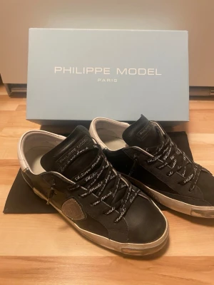Philippe Model svarta sneakers - Snygga svarta sneakers från Philippe Model med vita detaljer och logotyp på sidan. Skorna har svarta snören med text och en klassisk låg siluett. Ovandelen är i mocka och läder, och sulan är vit med slitna detaljer för en cool look.