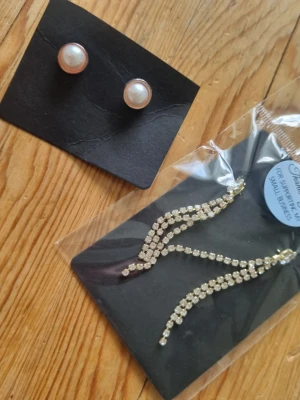 Två par örhängen, pärlor och strass - Säljer två par örhängen: ett par klassiska vita pärlörhängen med guldkant och ett par långa, glittrande örhängen med strass i vågig form. Perfekta för dig som vill ha både stilrent och extra bling i smyckeslådan.