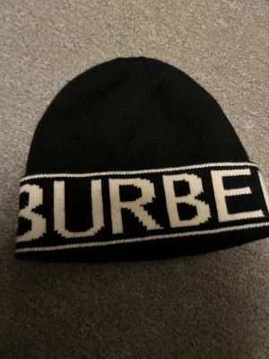 Svart mössa från Burbery - Snygg svart mössa från Burberry med vit logotyp i bred text runt kanten. Tillverkad i mjukt stickat material som håller dig varm och ger en clean look. Perfekt accessoar för dig som gillar klassisk design med en modern twist.