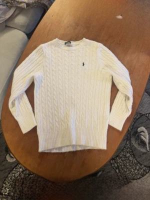Vit kabelstickad tröja från Polo Ralph Lauren - Kabelstickad vit tröja från Polo Ralph Lauren med rund halsringning och den klassiska lilla logotypen broderad på bröstet. Tröjan har långa ärmar och ribbade muddar vid ärmslut och nederkant. Perfekt för lager på lager under kyliga dagar.