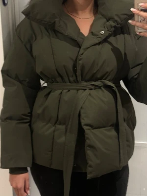 Olivgrön dunjacka från Copperose - Snygg olivgrön dunjacka från Copperose med bälte i midjan och hög krage. Jackan har en puffig siluett och stängs med knappar framtill. Perfekt för våren och ger en trendig look.