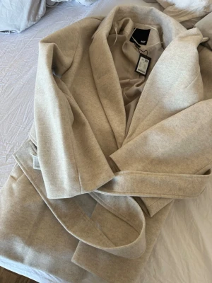 Beige kappa från 157 med skärp - Stilren beige kappa från 157 i mjukt material med klassisk krage och avtagbart midjeskärp. Kappan har en slät yta och helfodrad insida för extra komfort. Perfekt för dig som vill ha en enkel men snygg look till hösten eller våren.