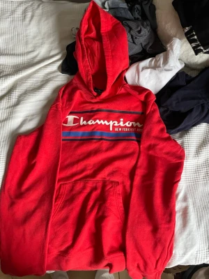 Röd hoodie från Champion med tryck - Snygg röd hoodie från Champion med stor logga och texten 'New York EST 1919' på bröstet. Hoodien har huva, känguruficka och ribbade muddar. Tillverkad i mjukt bomullsmaterial, perfekt för en avslappnad streetwear-look.
