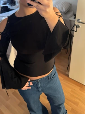 Svart topp med snörning och vida ärmar - Säljer en svart topp med unika detaljer som snörning längs ärmarna och öppna axlar. 
