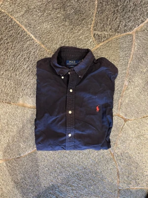Mörkblå skjorta från Polo Ralph Lauren - Klassisk mörkblå skjorta från Polo Ralph Lauren med knappslå och vit knappdetalj. Skjortan har lång ärm och ikonisk röd logobrodyr på bröstet. Nypris: 1449, mitt pris: 199