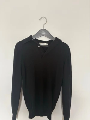 Svart pike i bomull  - Stilren svart tröja från Suitsupply med klassisk krage och knappdetalj vid halsen. Tröjan har långa ärmar och ribbade muddar vid ärmslut och nederkant. Perfekt för en clean och enkel look. Nypris 1299