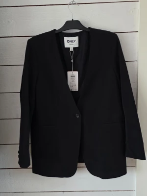 Svart kavaj från ONLY tailoring - Stilren svart kavaj från ONLY tailoring med enkelknäppning och klassisk passform. Kavajen har två fickor framtill och är tillverkad i ett mjukt, vävt material som ger en snygg siluett. Perfekt för att lyfta vilken outfit som helst.