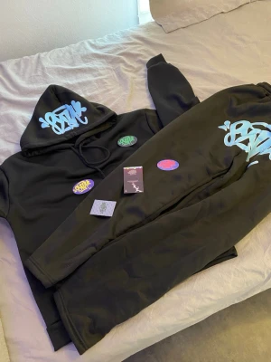 Syna world tracksuit - Säljer ett svart set med hoodie och byxor, båda med ljusblå graffiti-tryck. Hoodien har dragsko i huvan och ribbade muddar. Byxorna har samma tryck på ena benet. Setet är i mjukt material, troligen bomull eller bomullsblandning. Perfekt för en avslappnad streetwear-look. Den är velt ny och den har lapp med sig. Inte alls använda. Fin, bra och passar.