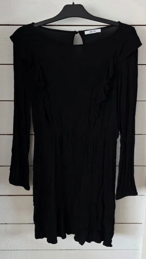 Svart volangklänning från About You - Snygg svart klänning från About You med volangdetaljer framtill och långa ärmar. Klänningen har en lätt rynkad struktur och en liten knapp i nacken. Perfekt för dig som gillar enkel men stilren design.