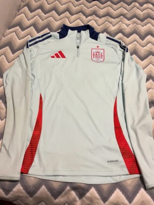 Ljusblå Spanien träningströja Adidas - Ljusblå långärmad träningströja från Adidas med Spaniens landslagsemblem på bröstet och röda detaljer. Tröjan har dragkedja vid halsen, mörkblå axelpartier och röda inslag på sidorna. Tillverkad i funktionsmaterialet Aeroready.
