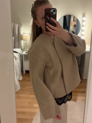 Beige vårjacka från Gina - Säljer en vårjacka med minimalistisk design och rundad halsringning. Jackan har vida ärmar och en rak, boxig passform utan synliga knappar eller dragkedja. Perfekt för dig som gillar stilrena och trendiga plagg.