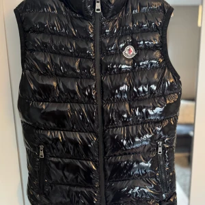 Svart dunväst märkt Moncler  - Snygg svart dunväst märkt Moncler i glansigt material med klassisk logga på bröstet. Västen har två dragkedjefickor framtill och quiltad design. Stängs med dragkedja hela vägen upp. OBS Västen har en fläck vid halsen som vad jag vet inte går bort men syns inte så mycket 