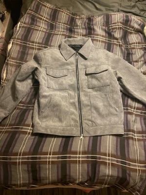 Grå skinnjacka med fickor - Snygg grå vårjacka med rak passform och två stora bröstfickor med lock samt sidofickor. Jackan har en klassisk krage och dragkedja framtill. Perfekt för dig som vill ha en stilren och enkel look.