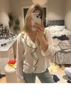 Vit blus med volang och svarta kanter - Säljer en vit blus med långa ärmar och volangdetalj framtill som ramas in av en svart kant. 
