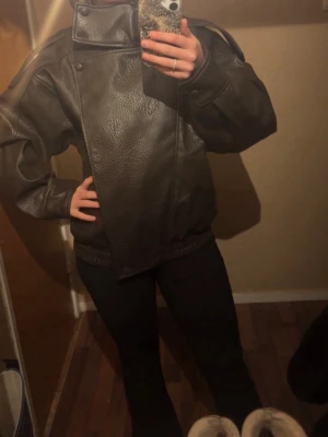 Mörkbrun oversized skinnjacka - Säljer en mörkbrun oversized skinnjacka med hög krage och breda axlar. Jackan har en unik asymmetrisk knäppning framtill och ribbade muddar i midjan. Perfekt för dig som gillar en cool och edgy stil.