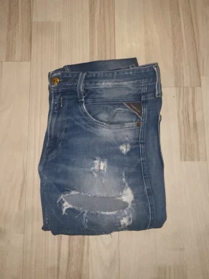 Blå slitna jeans från Replay - Säljer ett par blå jeans från Replay med snygga slitningar och distressed detaljer både fram och bak. Klassisk femficksmodell med läderpatch bak i midjan. Jeansen har en avslappnad passform och är tillverkade i denim.