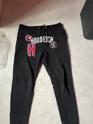 Svarta Hoodrich mjukisbyxor - Säljer ett par svarta mjukisbyxor från Hoodrich med vita och röda broderade detaljer och loggor framtill. Byxorna har dragsko i midjan och smal passform. Perfekta för chill dagar och streetwear-stil.