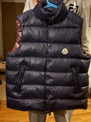 Mörkblå dunväst från Moncler - Snygg mörkblå dunväst från Moncler med hög krage och två dragkedjefickor. Västen har ett klassiskt quiltat mönster och Moncler-logga på bröstet. Insidan är vinröd och materialet är glansigt och fyllt med dun för extra värme.