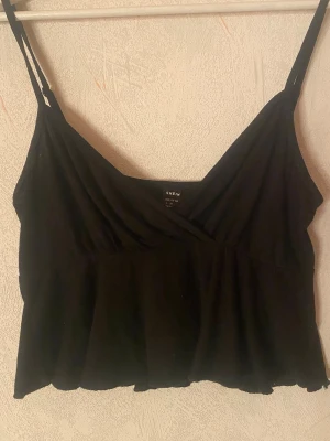 Svart linnetopp - Snygg svart linnetopp från SHEIN med smala axelband och omlott-detalj framtill. Toppen har en lätt utsvängd nederkant och är perfekt för dig som gillar enkel men trendig stil. Materialet känns mjukt och lätt.