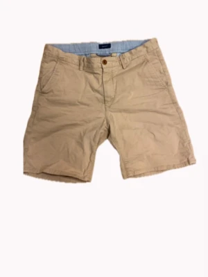 Beige shorts från Gant - Klassiska beige shorts från Gant med normal passform. Tillverkade i bomull med knappgylf och sidofickor. Perfekta för varma dagar och enkel att matcha med olika stilar.