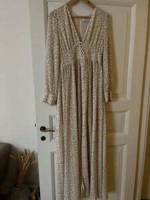 Blommig långklänning från Adoore - Långklänning från Adoore i ljusbeige med små gula blommor. Klänningen har v-ringning, knappar framtill, lång ärm och ett luftigt, flowy fall. Perfekt för sommarens alla tillfällen och ger en romantisk vibe.