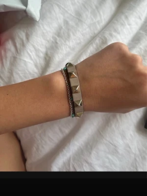 Rosa Valentino armband med nitar - Snyggt armband i  läder med stora, fyrkantiga guldfärgade nitar längs hela bandet. Nypris 2200kr ( kolla kvitto). Armbandet har en cool och edgy stil som passar perfekt till streetwear eller för att ge din outfit en tuff touch. Justerbart spänne för bästa passform. 