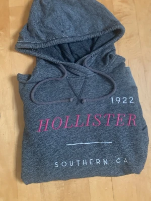 Grå hoodie från Hollister - Mysig grå hoodie från Hollister med tryck i vitt och rosa framtill. Klassisk huva med dragsko och mjukt material på insidan. Texten 'HOLLISTER 1922 SOUTHERN CA' ger en cool och avslappnad vibe. Perfekt för chill dagar eller när du vill ha en casual look.