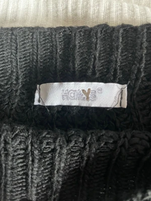 Svart stickad tröja från HOLY&S - Svart stickad tröja från HOLY&S med grov ribbstickning och rund hals. Tröjan har lång ärm och en avslappnad passform, perfekt för lager-på-lager-stil. Materialet är mjukt och ger en mysig känsla.