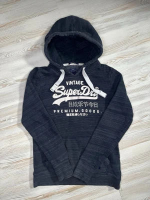 Mörkgrå Superdry hoodie med tryck - Snygg mörkgrå hoodie från Superdry med vit logga och text på bröstet🤩 Hoodien passar nån i Xs och är i fint skick🙌🏻 Hör av er vid funderingar samt snabbaffär😇